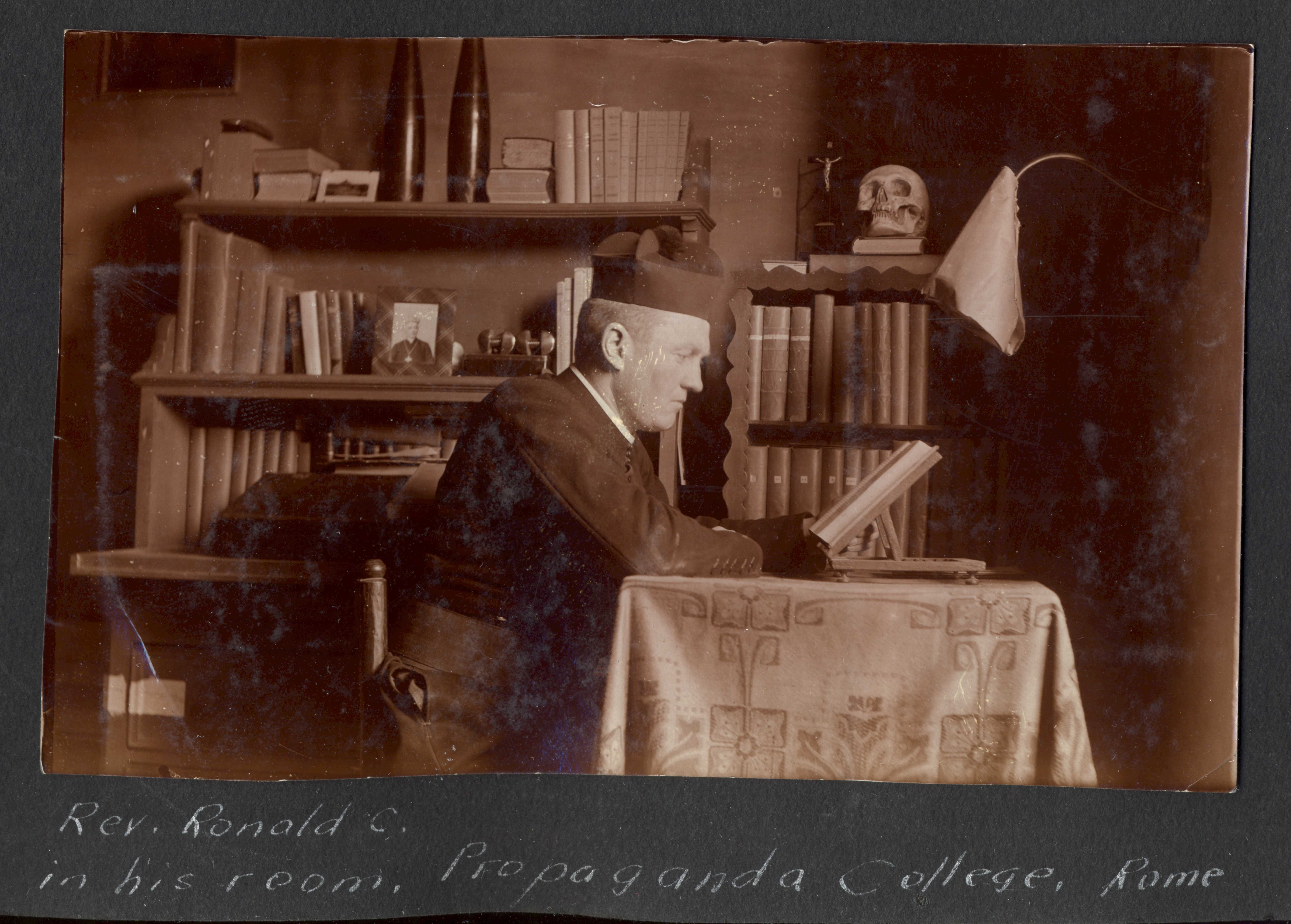 Photographie en noir et blanc – R.C. MacGillivray est montré de profil, en train de lire un livre, assis à une table couverte d’une longue nappe brodée. On aperçoit des étagères de livres derrière lui. Il porte un uniforme et un chapeau de couleur foncée.